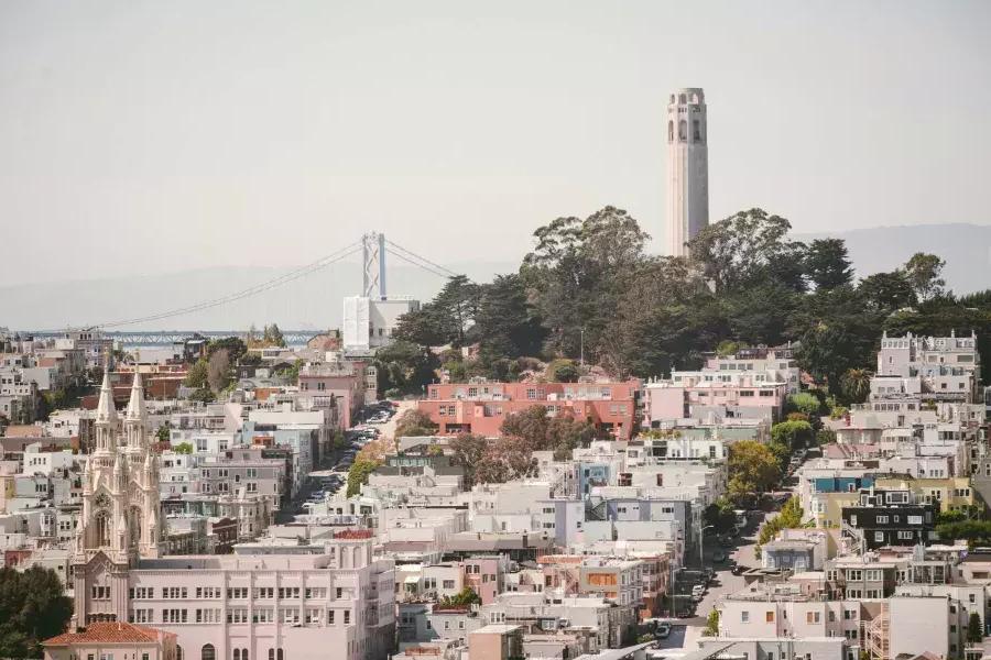 A Coit Tower de São Francisco é retratada 和 a Bay Bridge ao fundo e uma colina coberta de casas em primeiro plano.